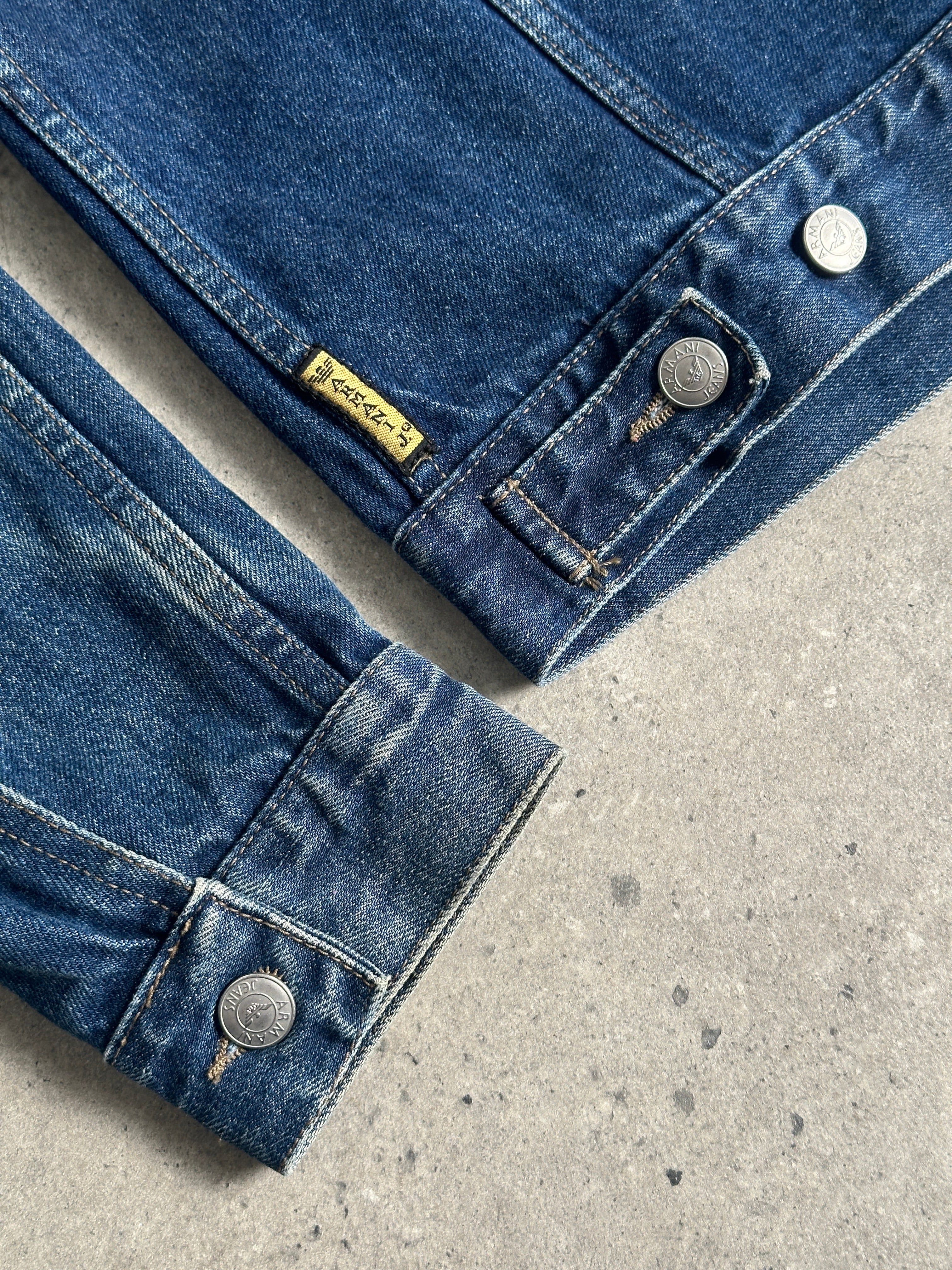 Armani Jeans mid wash denim jacket in size S/M displayed on a hanger, featuring classic stitching and button closures.