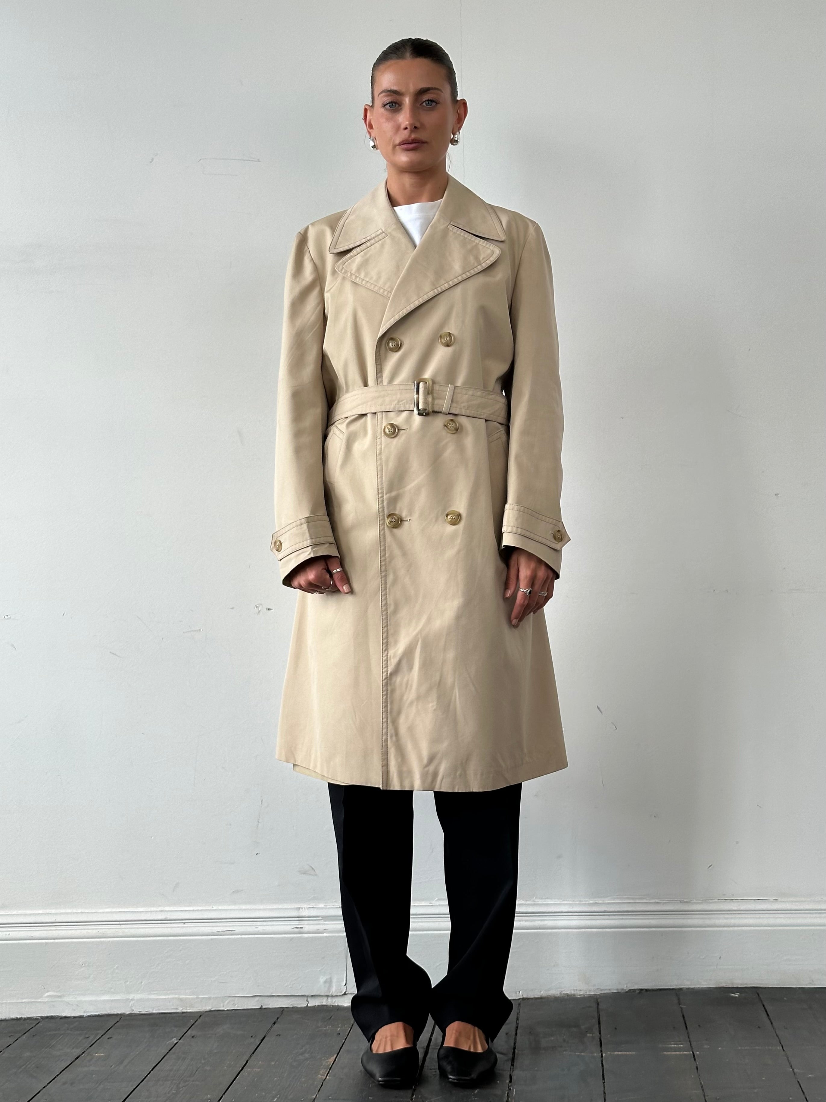 Vintage double-breasted belted trench coat in classic style, displayed on a hanger with visible fabric details and timeless design.